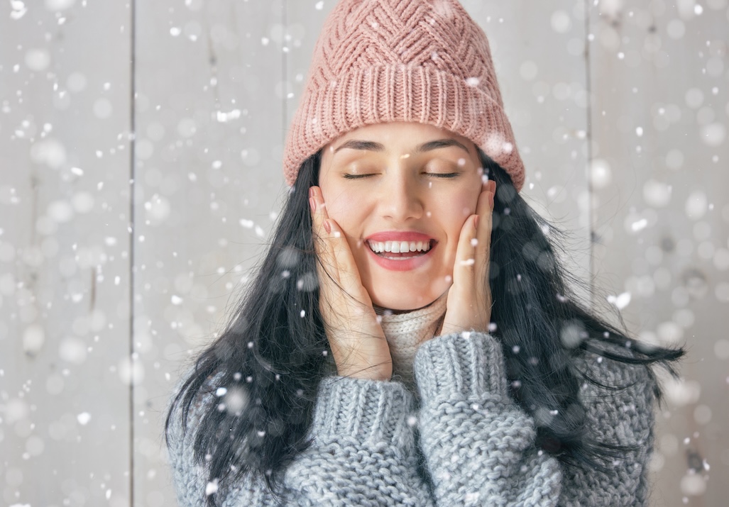 Smiling woman in winter hat with glowing skin in falling snow, representing holiday wellness and self-care in Nashville.