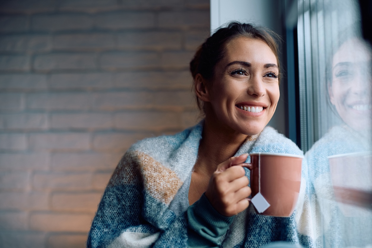 Smiling woman wrapped in a blanket sipping warm tea by a window.